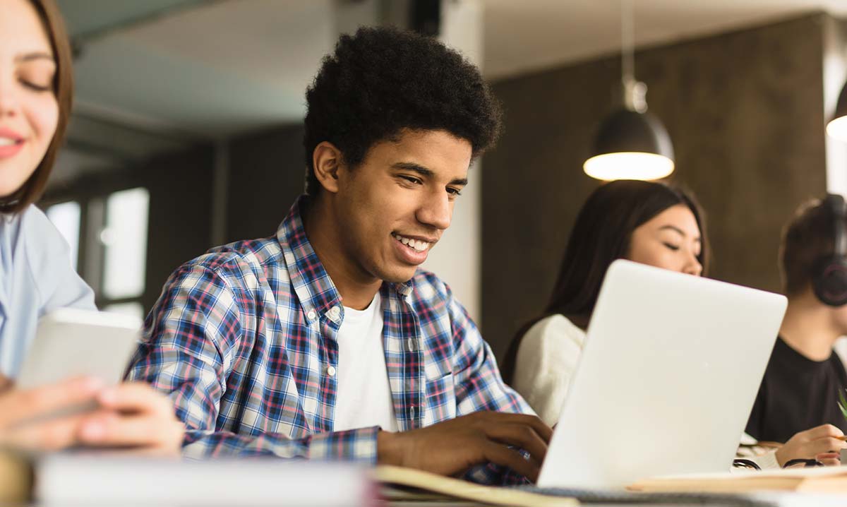 Ein Teenager sitzt mit weiteren Personen nebeneinander an einem Tisch und blickt freundlich auf seinen aufgeklappten Laptop.