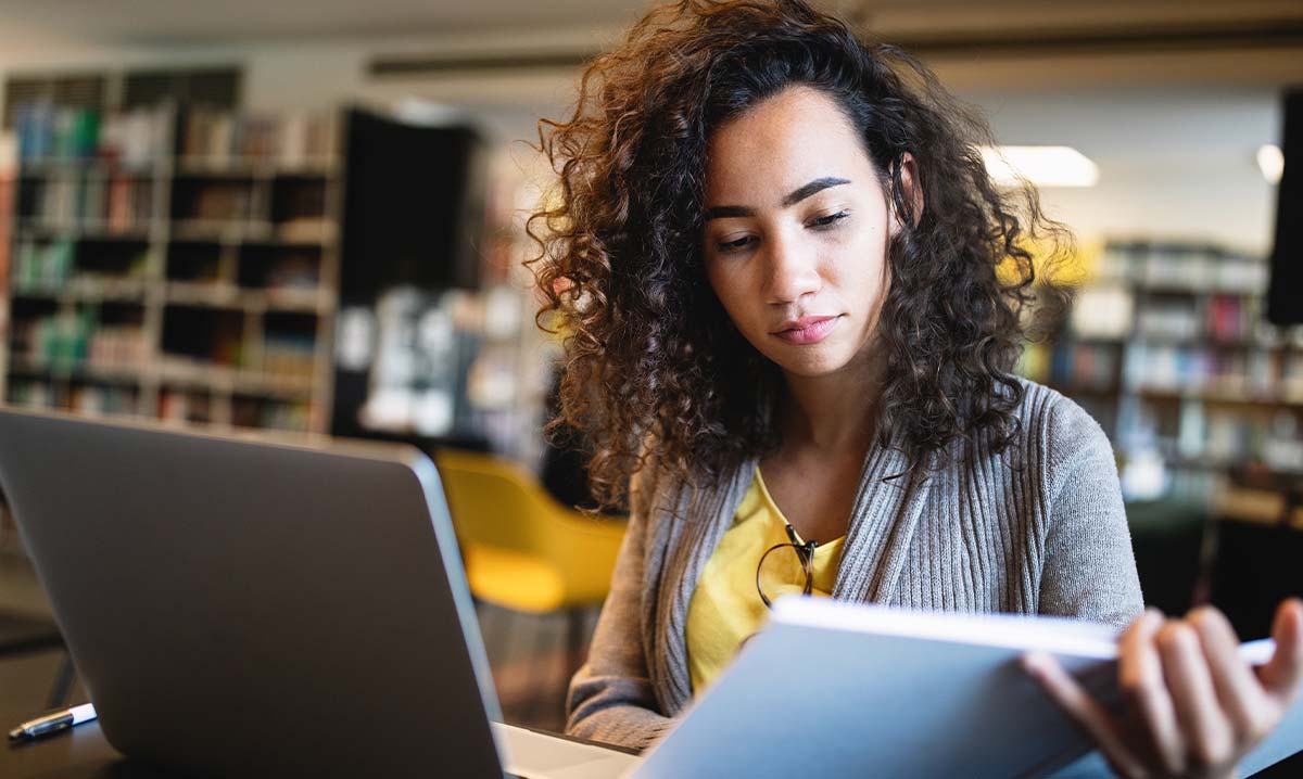 Eine junge Frau sitzt in einer Bibliothek an einem Arbeitsplatz mit aufgeklappten Laptop und blickt konzentriert in ein aufgeschlagenes Buch.